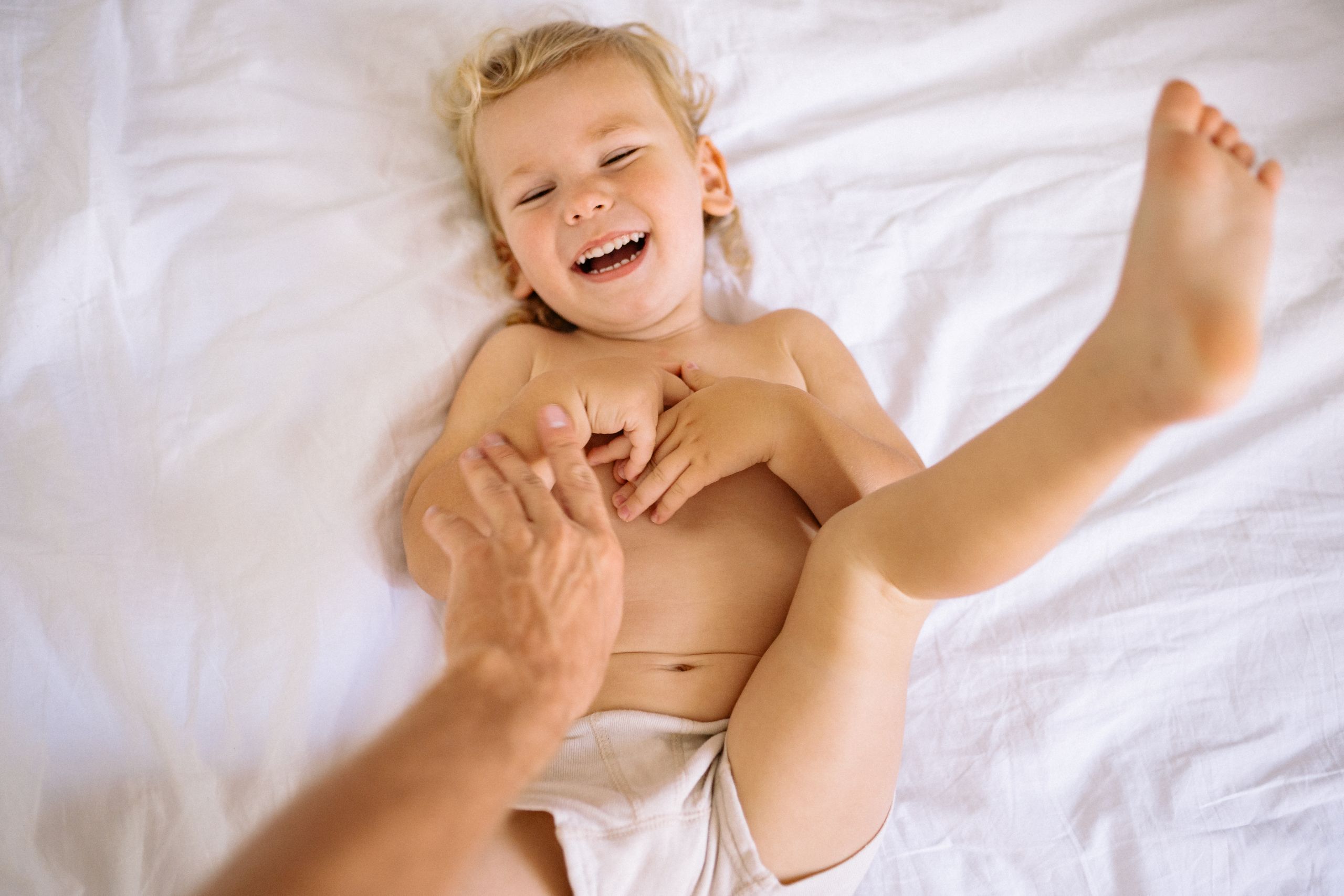 A happy child laughing while being tickled in a playful moment on a white bed.